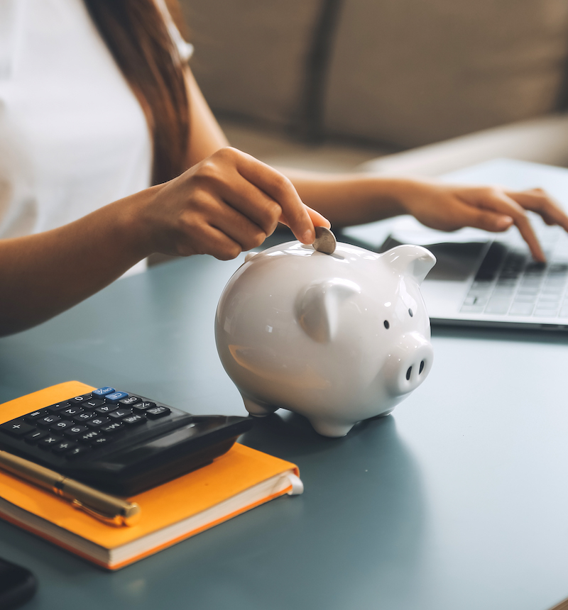 Young Asian woman saving for retirement Saving money through a piggy bank and taking notes on notebook, savings concept.